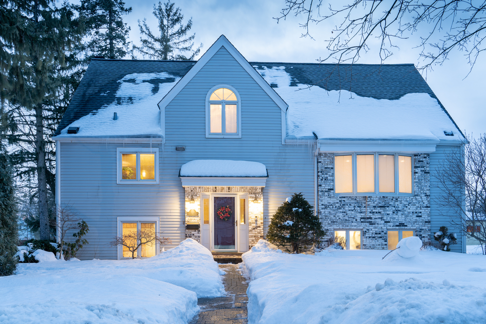 white house with stone detailing on the front siding exterior in the winter with snow on the black shingled roof and lights on inside while the storm finishes passing falling down over the town